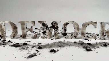 Yavaş çekim, kömürleşmiş ahşap harfleri gösteriyor. Demokrasi kelimesini heceliyor, küllerin arasında beyaz, pürüzsüz bir arkaplan karşısında oturuyor. Demokrasinin saldırı altında olduğunu gösteriyor.. 