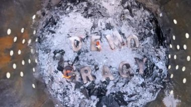 Yavaş çekim, kömürleşmiş ahşap harfleri gösteriyor. Demokrasi kelimesini heceliyorlar. Çöp tenekesinde kül yığınının içinde yanarak, demokrasinin tehdit edildiğini öne sürüyorlar..