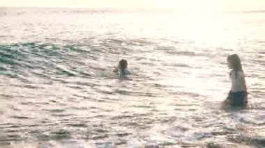 Asian children indulge and play in the water. Little girls and a boy are waiting for the waves, standing in the ocean and enjoying their childhood at sunset. Bali, Indonesia, June 28, 2022