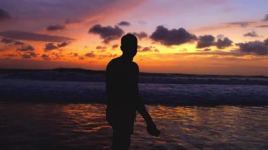 Gün batımında sahilde yürüyen bir adamın silueti. Tatillerde tropik adalarda dinleniyor, rahat bir hayat sürüyor. İnsan, okyanus boyunca bir akşam yürüyüşü boyunca güzel, sıcak bir günbatımının tadını çıkarır..