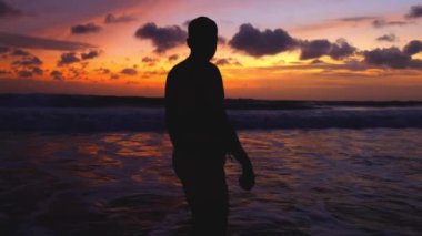 Okyanus boyunca yürüyen bir adamın silueti parlak, çok renkli bir günbatımının arka planına karşı. Dalgaların huzur ve sesinin tadını çıkaran bir adam. Güzel bir manzaranın yavaş çekim 4k görüntüsü