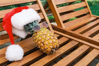 Güneş gözlüğü takan komik ananasın yan görüntüsü ve Noel Baba şapkası tropiklerde güneşli bir havada tahta güneş yatağında yatıyor. Tropiklerde Noel. Yazın kış tatili.