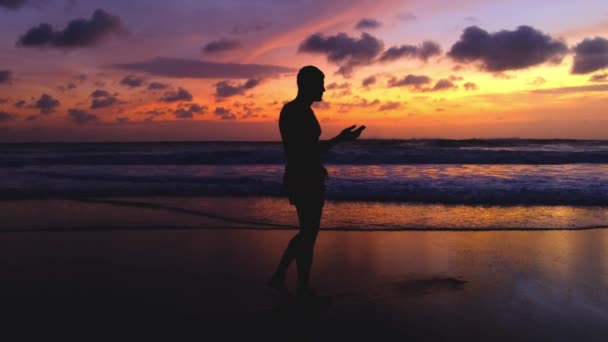シルエットの男の燃えるような色の夕日に対してビーチに沿って 携帯電話を探して歩く スローモーション美しい風景と人の4K映像 海を歩いている間に携帯電話を使う — ストック動画