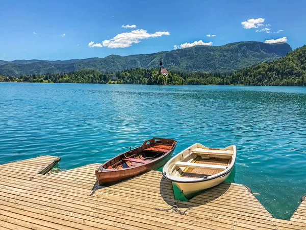 礼拝堂を背景に木製の桟橋で2隻の木製のボートと晴れた日にスロベニアのブレッド湖の森の丘 — ストック写真