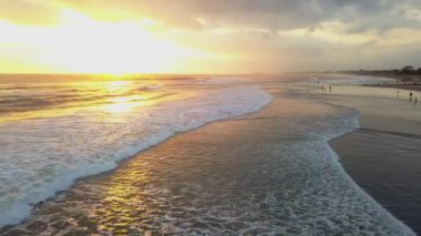 Hava aracı, gün batımında ıssız bir plajın üzerinden alçaktan uçuyor. Yuvarlanan dalgalarla kumsal kıyısı ve ufukta batan güneş. Deniz Burnu.