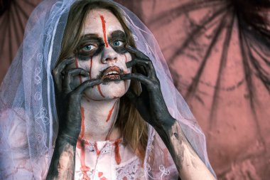 Duvağı ve sahne makyajıyla kameraya bakan zombi kadın portresi. Kız yüzüne beyaz makyaj ve kan lekeleri sürmüş.
