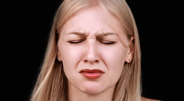 Headshot of a young crying woman experiencing grief or loss. Grimace of mental pain and closed eyes. Woman on black background. Girl is under severe stress and cannot restrain her emotions.