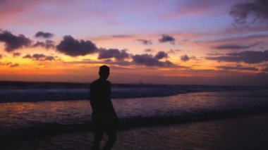 Gün batımında sahilde yürüyen bir adamın silueti. Tatillerde Bali adasında dinleniyor. Rahat bir yaşam tarzı. Adam, okyanus boyunca bir akşam yürüyüşü sırasında güzel, sıcak bir günbatımının keyfini çıkarıyor, 4k