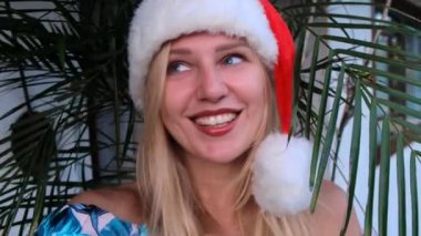 Noel Baba şapkalı genç ve güzel bir kızın portresi tatilde Noel 'i kutluyor, bitkilerin arka planındaki kameraya bakıyor. Çekici kadının bir video görüşmesi var ve konuşuyor.