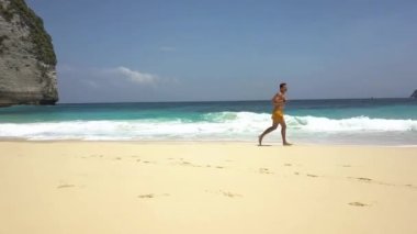 Genç ve mutlu atletik gezgin, turkuaz su ve beyaz köpükle tropikal plajın kumlu sahillerinde koşuyor. Kelingking Sahili, Nusa Penida, Endonezya. Seyahat, boş zaman, tatil