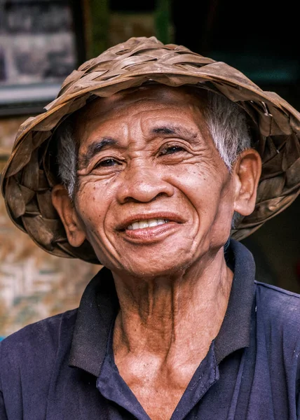 Vertical portrait of a South Asian Balinese senior man wearing a traditional Balinese cone hat. Elderly man looking at the camera smiling.