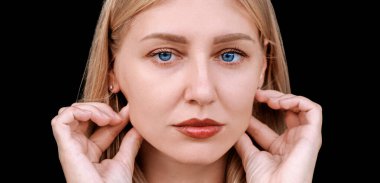 Plastic surgery, facelift, rejuvenation. Portrait of a beautiful middle aged woman looking at the camera, touching her cheekbones with her hands. Blue-eyed blonde caucasian girl on a dark background