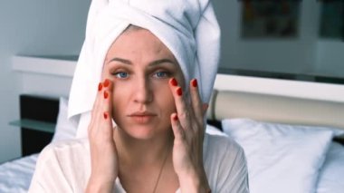 Portrait of a young woman looking at the camera, touching her face, wearing a towel on her head and a bathrobe. The concept of face and body care, beauty and anti-aging treatments.