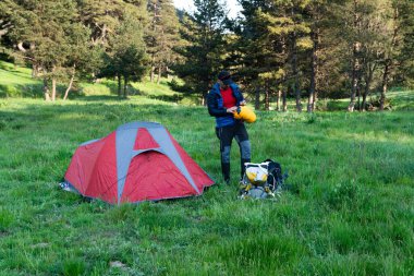 Ağaçların arasındaki çimlerin üzerinde kırmızı kamp çadırı ve sırt çantasıyla tanınan bir adam.