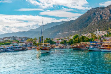 Kas, Antalya, Türkiye - 7 Ağustos 2024: Deniz kenarında demirlemiş ve yolcu bekleyen günlük tekne turları. Kas limanı ve Kas şehri ve arka planda dağ.