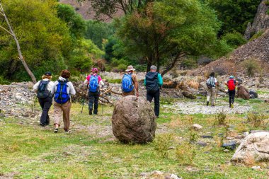 Bir grup yürüyüşçü sırık ve sırt çantalarıyla ormanlık ve kayalık bir vadide yürüyor..