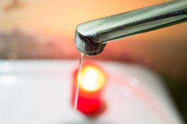 Yanan bir mumun arka planına karşı musluktan gelen ince bir su püskürtüsü. Savaş nedeniyle Ukrayna 'da elektrik ve su kesintileri