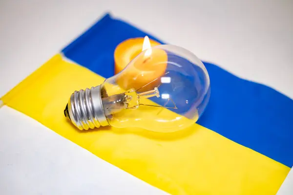 stock image Light bulb on the background of a burning candle and the Ukrainian flag. Blackout due to the war in Ukraine