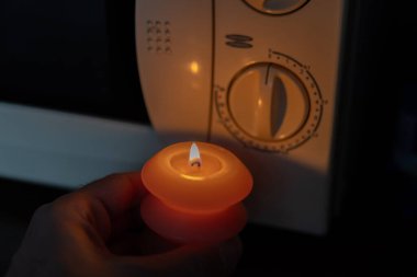 Elinde yanan mum, karanlıkta mikrodalga fırının yanında. Ukrayna 'da savaş nedeniyle elektrik kesintisi