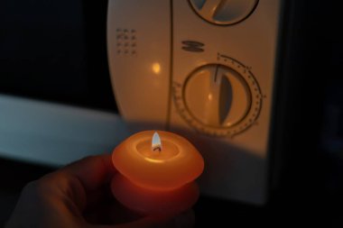 Burning candle in hand near microwave oven in dark. Power outage due to war in Ukraine clipart