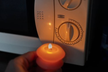 Burning candle in hand near microwave oven in dark. Power outage due to war in Ukraine clipart