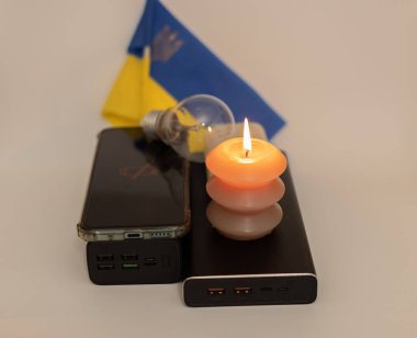 Elektrik bankalarının yanında yanan bir mum, boşaltılmış bir telefon ve Ukrayna bayrağının arka planında kırık bir ampul. Ukrayna 'daki savaş nedeniyle elektrik kesintileri