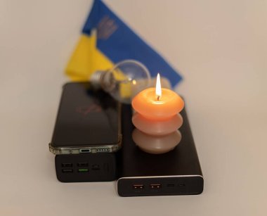 Elektrik bankalarının yanında yanan bir mum, boşaltılmış bir telefon ve Ukrayna bayrağının arka planında kırık bir ampul. Ukrayna 'daki savaş nedeniyle elektrik kesintileri