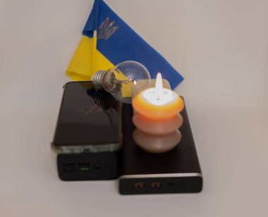 Elektrik bankalarının yanında yanan bir mum, boşaltılmış bir telefon ve Ukrayna bayrağının arka planında kırık bir ampul. Ukrayna 'daki savaş nedeniyle elektrik kesintileri