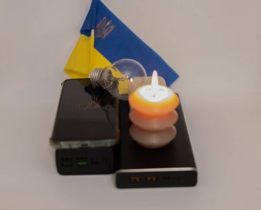 Elektrik bankalarının yanında yanan bir mum, boşaltılmış bir telefon ve Ukrayna bayrağının arka planında kırık bir ampul. Ukrayna 'daki savaş nedeniyle elektrik kesintileri