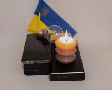 Elektrik bankalarının yanında yanan bir mum, boşaltılmış bir telefon ve Ukrayna bayrağının arka planında kırık bir ampul. Ukrayna 'daki savaş nedeniyle elektrik kesintileri