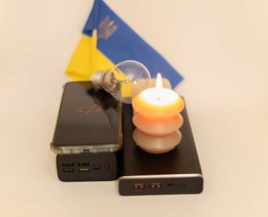 Elektrik bankalarının yanında yanan bir mum, boşaltılmış bir telefon ve Ukrayna bayrağının arka planında kırık bir ampul. Ukrayna 'daki savaş nedeniyle elektrik kesintileri