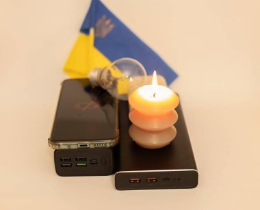 Elektrik bankalarının yanında yanan bir mum, boşaltılmış bir telefon ve Ukrayna bayrağının arka planında kırık bir ampul. Ukrayna 'daki savaş nedeniyle elektrik kesintileri