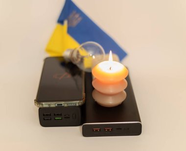 Elektrik bankalarının yanında yanan bir mum, boşaltılmış bir telefon ve Ukrayna bayrağının arka planında kırık bir ampul. Ukrayna 'daki savaş nedeniyle elektrik kesintileri