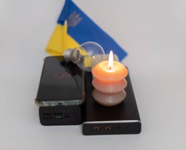 Elektrik bankalarının yanında yanan bir mum, boşaltılmış bir telefon ve Ukrayna bayrağının arka planında kırık bir ampul. Ukrayna 'daki savaş nedeniyle elektrik kesintileri