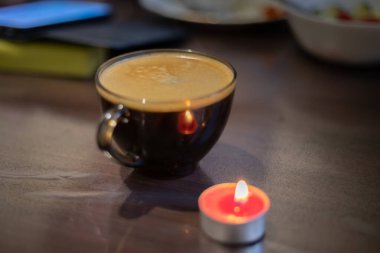Şeffaf bir fincanda kahve ve yakınlarda yanan bir mum. Elektriksel sorunlar nedeniyle karartma