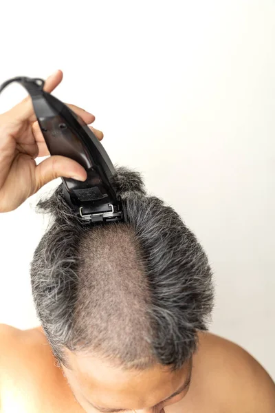 Handsome gray-haired man is cutting hair himself with an electric hair clipper.