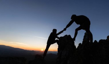 İki dağcı arasındaki yardım eli silueti. Yürüyüş yapan çiftler, dağlarda gün ışığıyla siluet oluşturmaya yardım ediyorlar. İnsanları yüksek uçurumlardan çıkarmaya yardım eden adamlar.