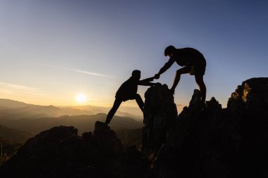 Siluet takım çalışması, dağ yamacına tırmanan erkek yürüyüşçüler ve bir tanesi yardım eli uzatıyor. Yardım eden insanlar ve takım çalışması konsepti.