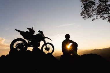 Motosikletli bir adam güzel ışıklara karşı, motosikletli bir adamın silüeti kayanın tepesinde, gün batımında, dayanıklı motosiklet yolculuğu konsepti..