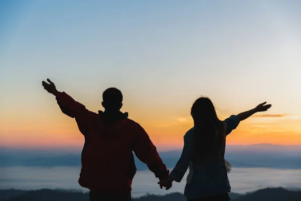 Silhouette of a romantic couple holding hands on top of a mountain at sunrise.