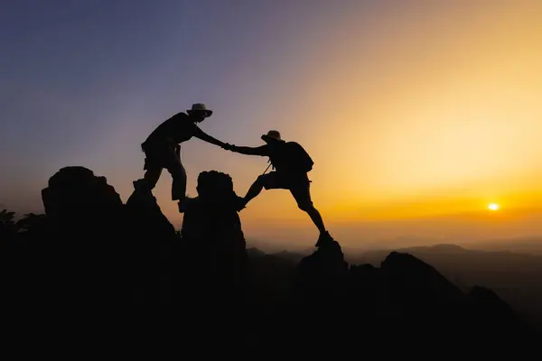 Silhouettes of two people climbing on mountain and helping. Teamwork of two men hiker helping each other on top of mountain climbing team beautiful sunrise. Help and assistance concept.