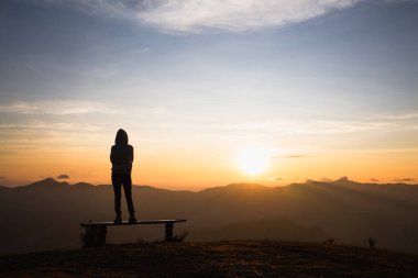 Bir dağın tepesinde tek başına durup güneşin doğuşunu izleyen bir kadının silueti..