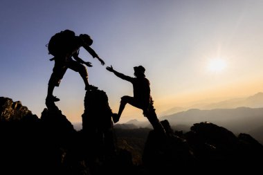 İki dağcının silueti vadinin yukarısındaki bir kayanın tepesinde yardım eli uzatıyor. Takım çalışması, iş birliği.