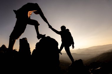 Yürüyüşçülerin silueti gün batımında bir dağa tırmanırken birbirlerine yardım ediyorlar. Yardım eden insanlar ve takım çalışması konsepti.