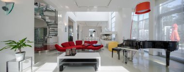 Lüks özel evin modern iç mimarisi. Geniş oturma odası, kırmızı kanepe, piyano, beyaz masa. Metal ve cam spiral merdiven.