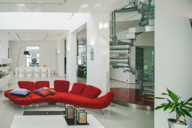 Lüks özel evin modern iç mimarisi. Geniş oturma odası, kırmızı kanepe, beyaz masa. Metal ve cam spiral merdiven.