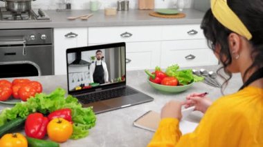 Bilgisayar ekranındaki erkek yemek blogcusu ev hanımlarına online video kameralı laptop öğretiyor, domates dilimliyor. Mutfaktaki kadın uzaktan aşçılık dersi alıyor. Şef öğretmeni dinle, defterine yazıyor.