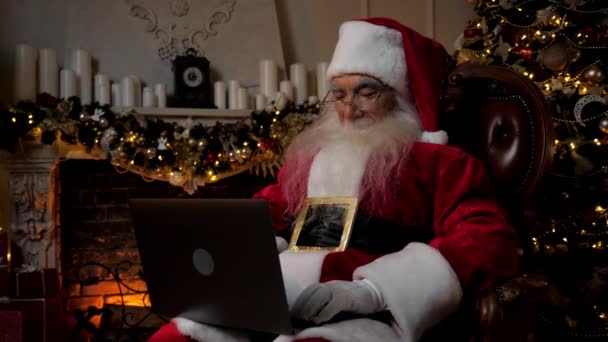 现代圣诞老人用笔记本电脑把头转过来 微笑着看着摄像机 积极的老祖父圣诞老人穿着圣诞老人的服装坐在椅子上 在电脑前工作 新年快乐 — 图库视频影像