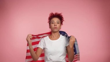 Ciddi Afro-Amerikan vatansever, kızıl saçlı, kıvırcık saçlı kadın Amerikan bayrağını omuzlarına asıyor. Amerikan bayrağıyla izole edilmiş pembe arka planda duran portre kadın. 4 Temmuz Amerika Bağımsızlık Günü