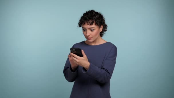 Shocked Curly Woman Using Smartphone Typing Browsing Isolated Light Blue — Stock Video